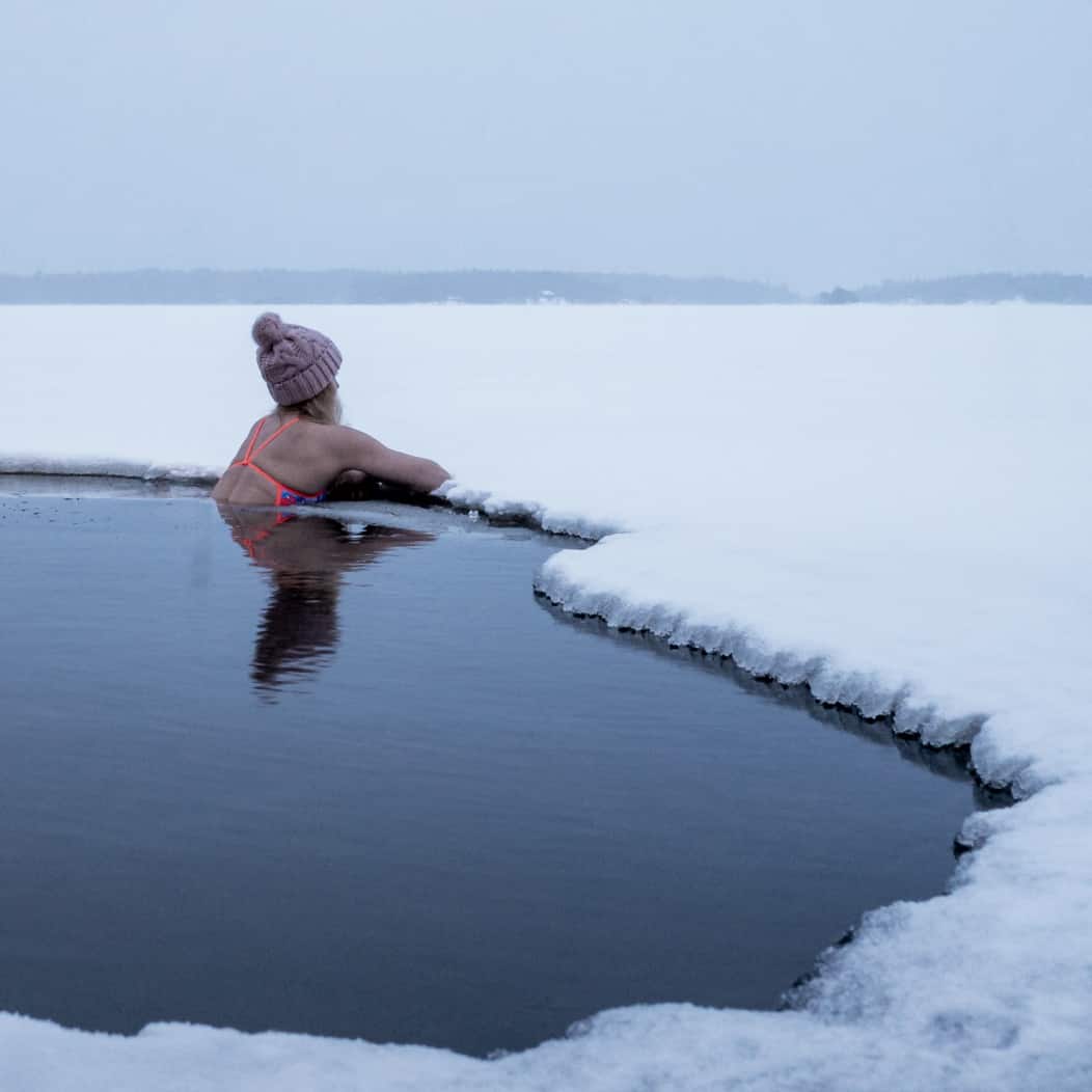 A woman wearing a warm hat sitting in a cold icy water hole, practicing cold therapy and breathwork in Bend, OR.