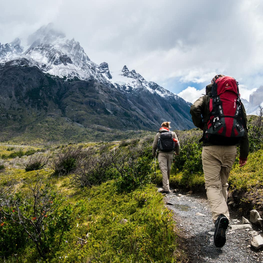 A couple carrying backpacks hiking with mountains in the background.